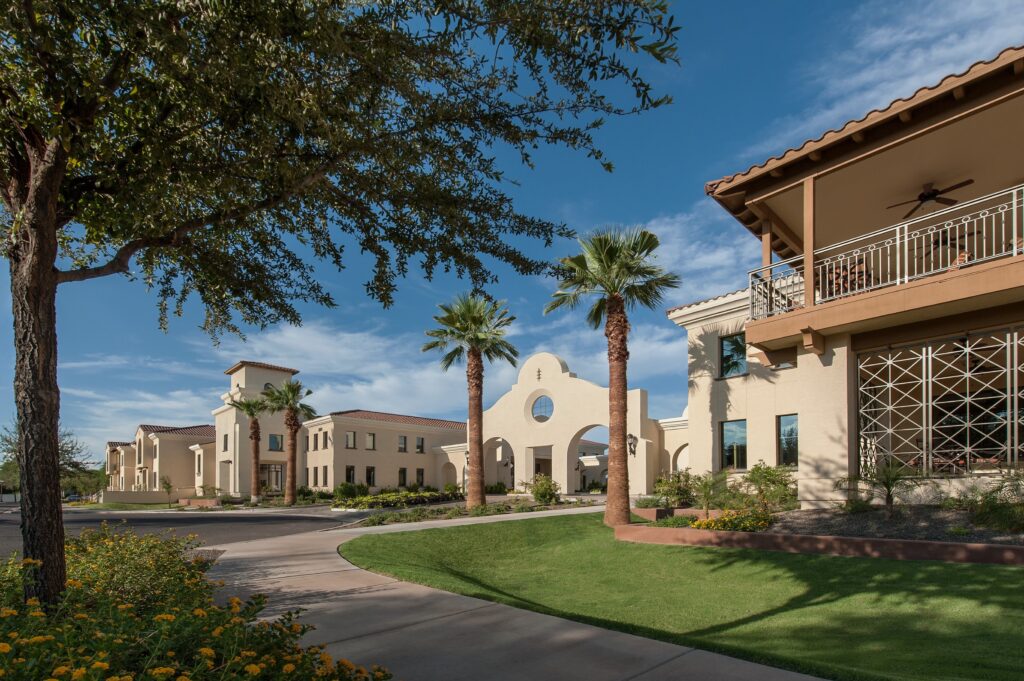 A Mediterranean-style building complex with arches and balconies, flanked by palm trees and landscaped gardens under a clear blue sky.