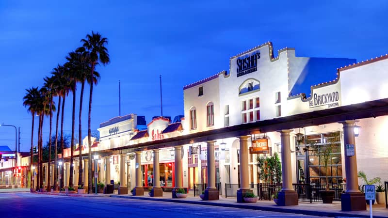 Street view of historic buildings at twilight, with palm trees lining the sidewalk. The facades are illuminated by lights, with visible signs and shop entrances.