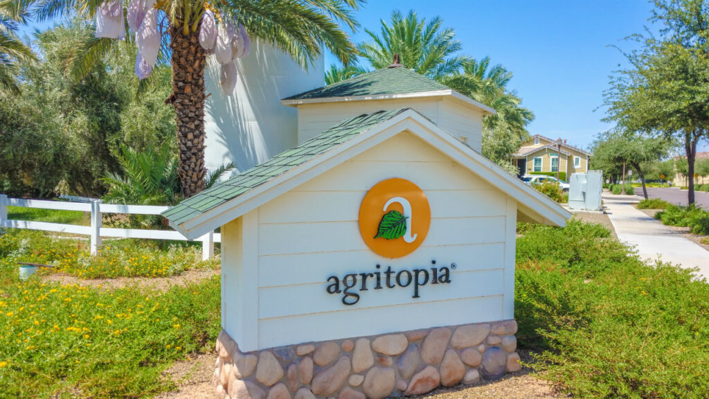 Sign reading "Agritopia" with a leaf logo, set against a backdrop of palm trees and a clear blue sky.