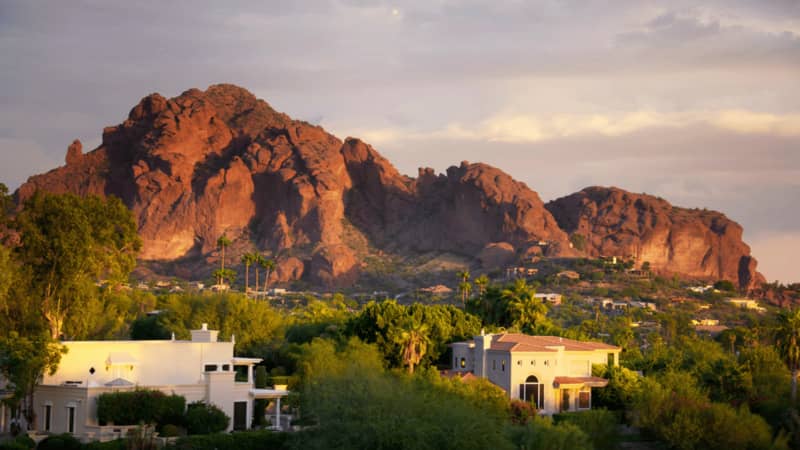 Camelback Mountain at sunset with a few houses in the foreground, surrounded by green trees and shrubs.