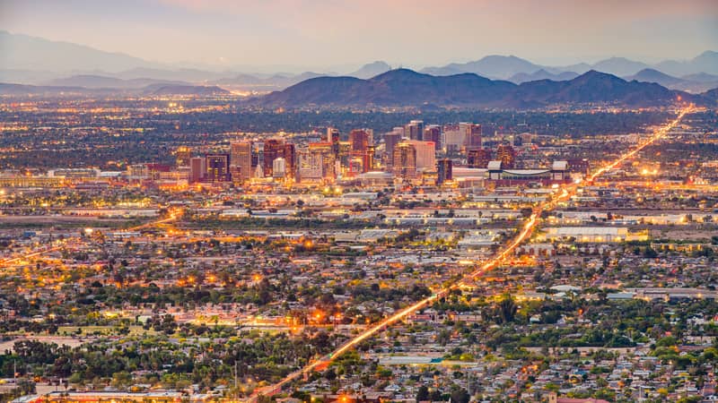 Aerial view of a cityscape at sunset, featuring a downtown area with tall buildings, surrounding residential areas, and distant mountains under a hazy sky.
