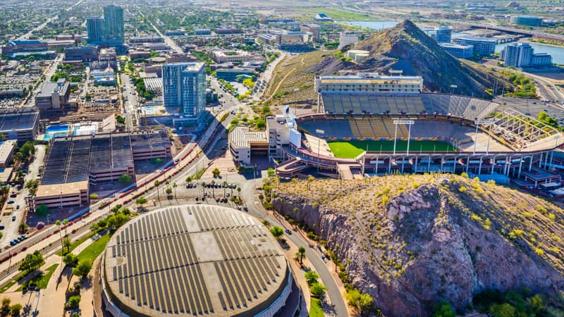 Aerial view of a cityscape featuring a large circular building, a stadium, a prominent hill, and various urban structures amidst a sprawling city environment.