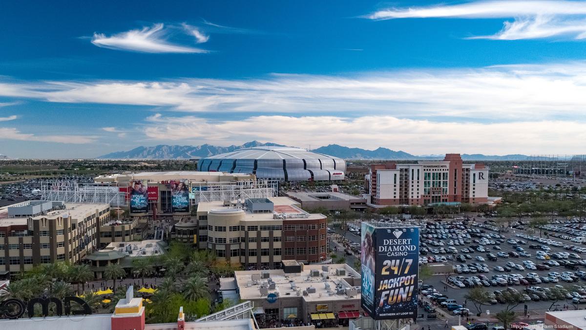 Aerial view of a large stadium surrounded by parking lots and adjacent buildings. Mountains are visible in the distance under a blue sky with scattered clouds.