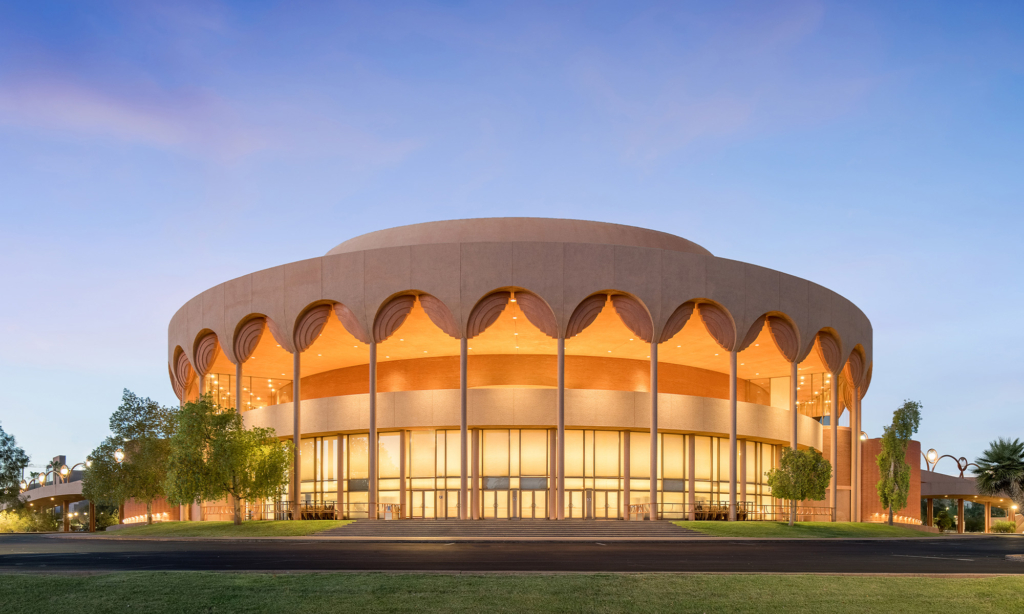Round modern building with a scalloped roofline and large windows, surrounded by trees, under a clear sky.