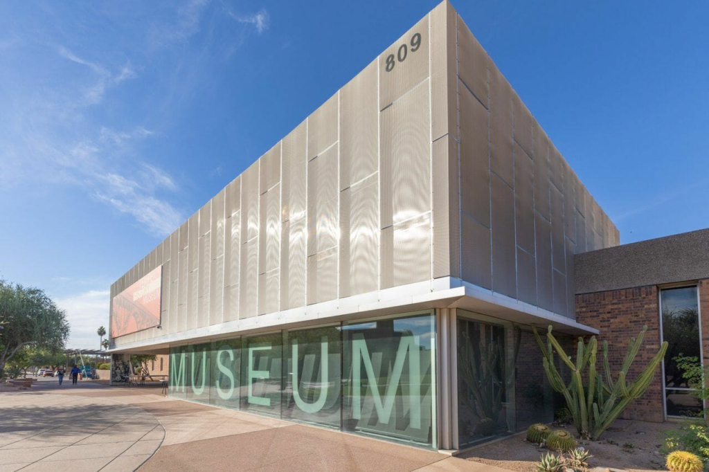 Contemporary building with large windows displaying "MUSEUM" and an address number "809" on the upper corner. Clear sky and surrounding cacti are visible.