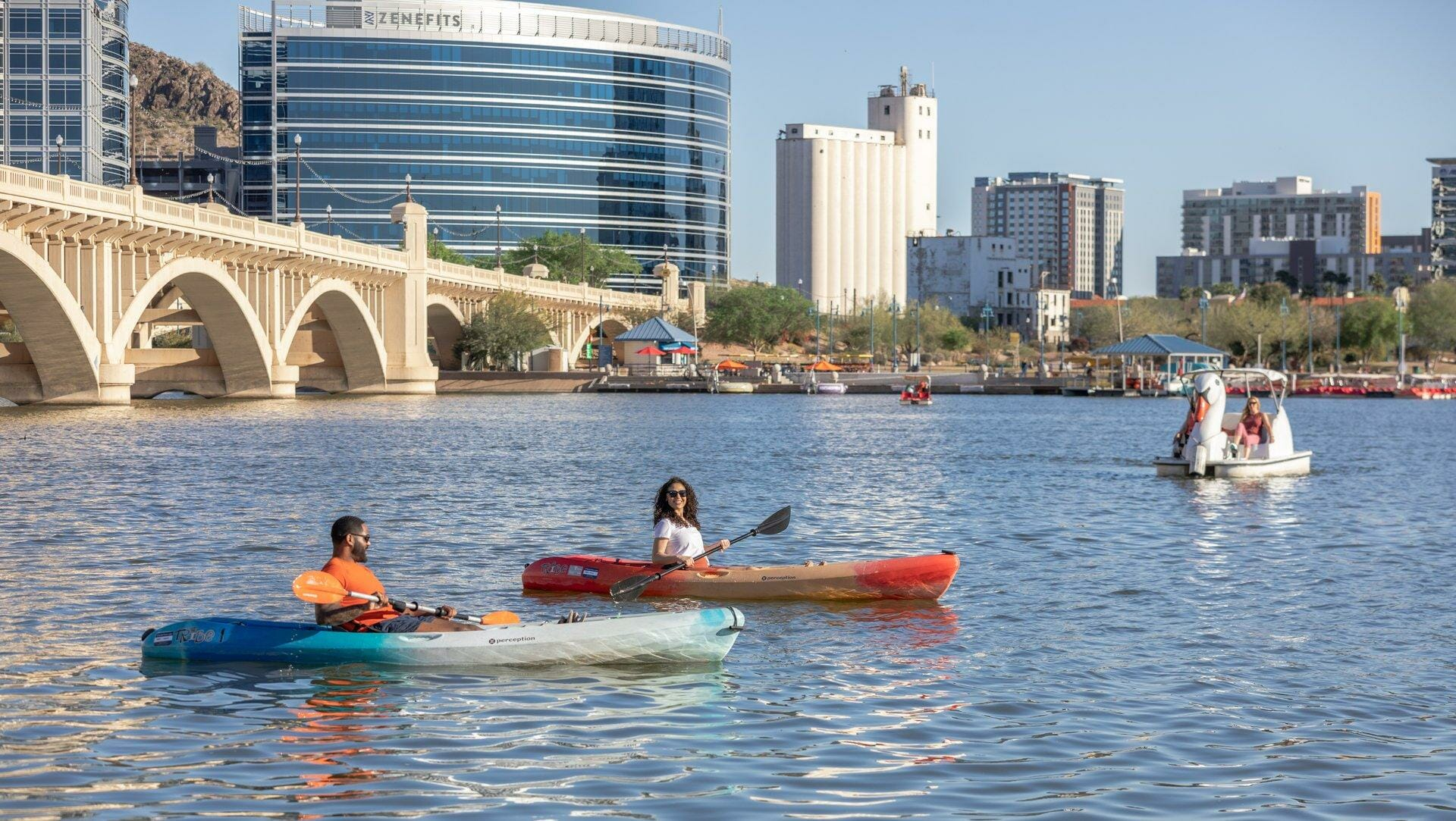 Two people kayaking on a calm river near an arched bridge, with a cityscape and several buildings in the background under a clear sky.