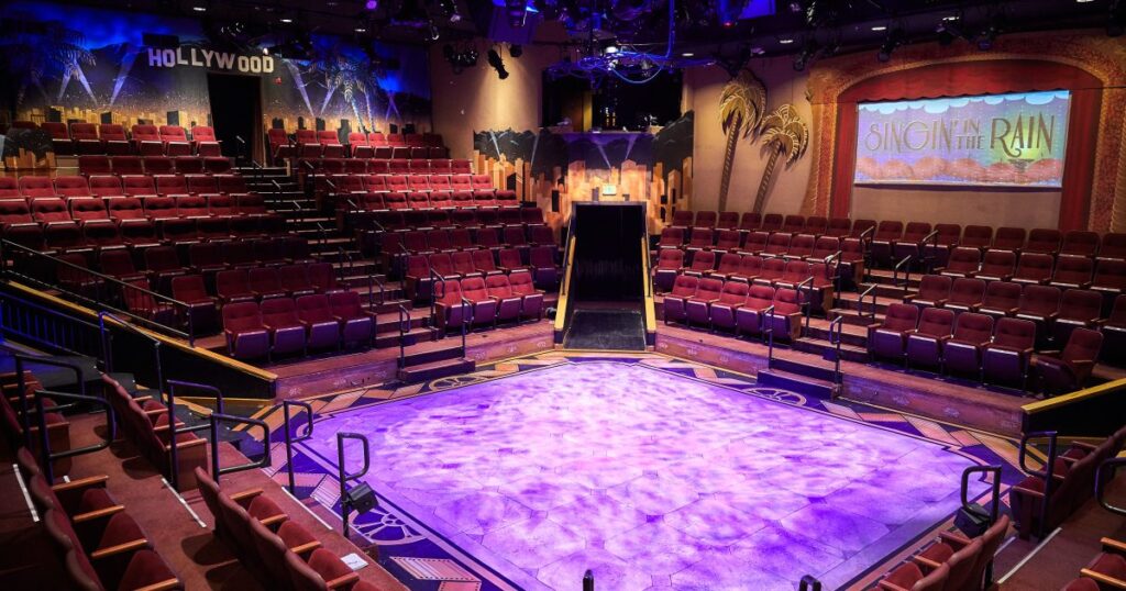 An empty theater with a purple-lit stage, rows of red seats surrounding it, and "Hollywood" and "Singin' in the Rain" signs at the back.