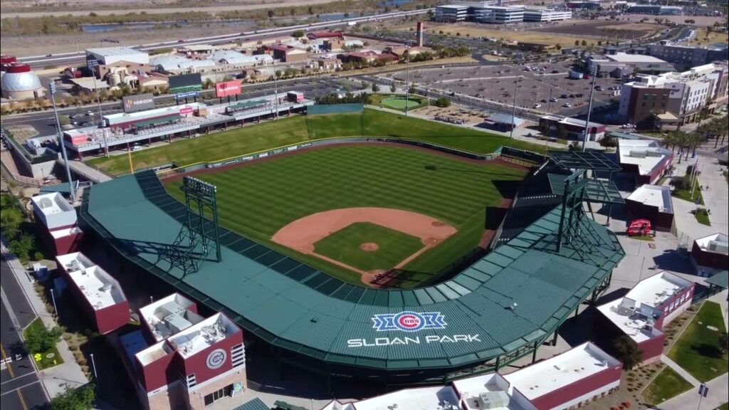 Aerial view of a baseball stadium with green seating and a field. The roof reads "Sloan Park." Surrounding areas include buildings, roads, and parking lots.